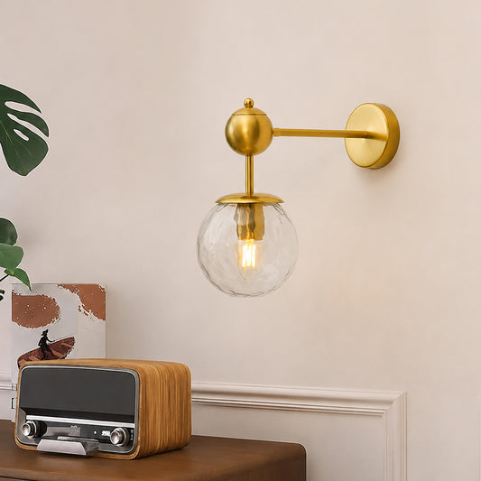 A modern wall-mounted sconce with a brushed gold metal arm and a clear glass globe shade, shown lit in a styled interior setting with a wooden table, vintage radio, and plant leaves.
