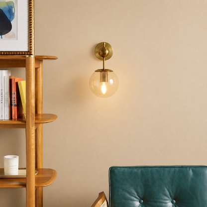 A brass wall sconce with a clear glass globe shade glowing with warm light, mounted on a beige wall in a styled interior next to a wooden shelf and a teal chair.