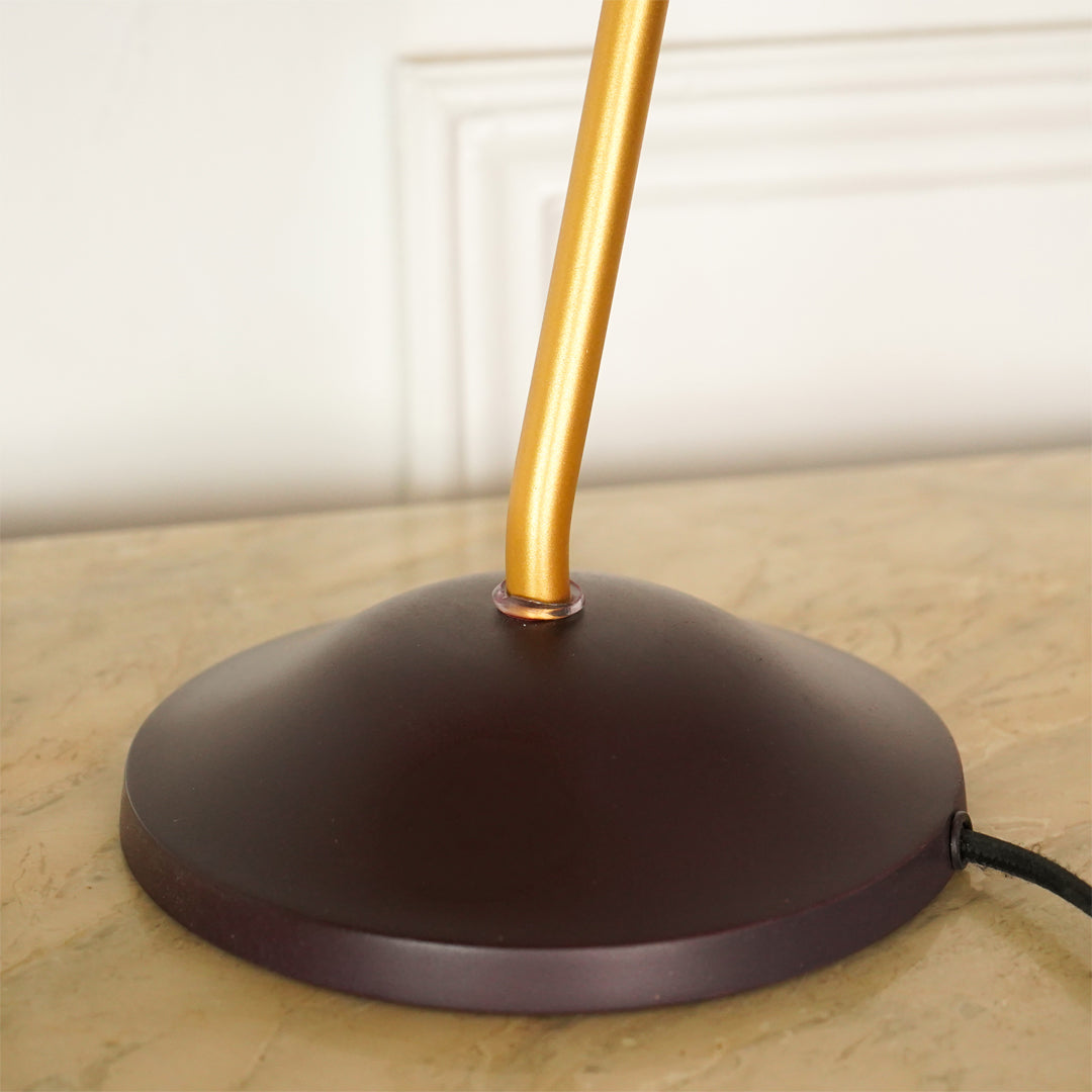 A close-up of a table lamp base with a dark brown dome-shaped metal shade and a gold-finished vertical stem, resting on a beige stone surface against a white paneled wall.