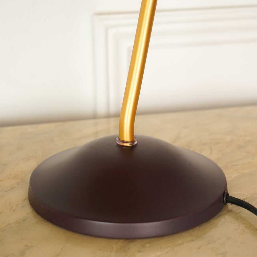 A close-up of a table lamp base with a dark brown dome-shaped metal shade and a gold-finished vertical stem, resting on a beige stone surface against a white paneled wall.