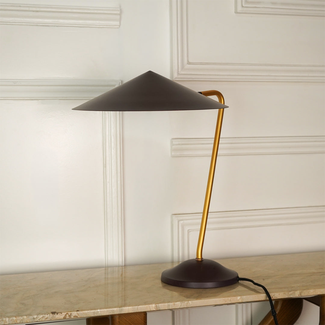 A modern table lamp with a dark brown conical shade and base, and a curved gold metal stem, shown on a marble tabletop against a white paneled wall.