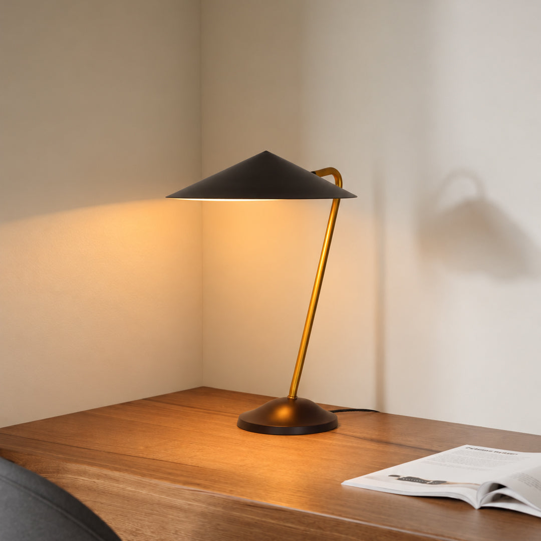 A modern table lamp with a matte black conical shade and a warm brass-colored curved stem, illuminated on a wooden desk against a beige wall.