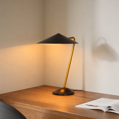 A modern table lamp with a matte black conical shade and a warm brass-colored curved stem, illuminated on a wooden desk against a beige wall.