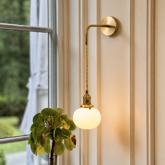 A wall-mounted brass sconce with a ribbed glass globe shade, glowing warm white, mounted on a cream paneled wall next to a window with a green plant in the foreground.