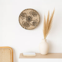 A round woven decor hangs on a white wall above a wooden shelf with a white vase of tall dried grass and a notebook near a cane chair.