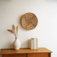 A round woven wall decor hangs above a wooden cabinet displaying a white vase with dried grass and a row of books against a white wall.