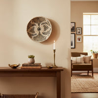 A round woven basket hangs on a beige wall above a wooden table with a candle and books next to a view of a sitting area with a brown chair.