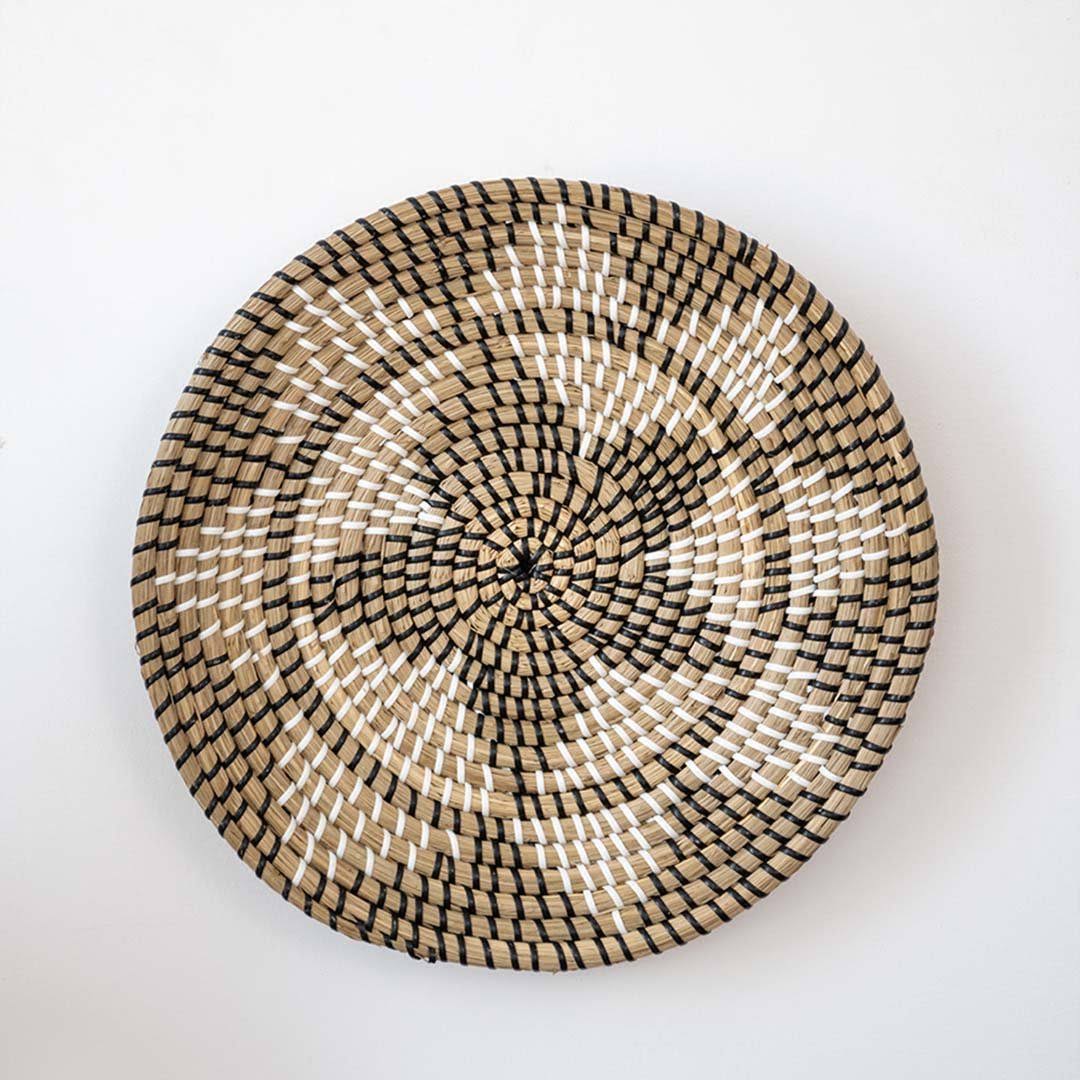 Handwoven seagrass round wall basket decor in natural beige and black with geometric floral woven pattern on white background.