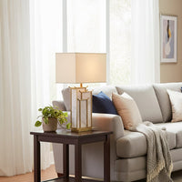 White shade gold and glass table lamp on wooden side table with light off beside sofa for modern bright décor styling.