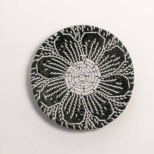 Round handcrafted black cream woven seagrass wall basket with detailed multi petal flower pattern on white background image.