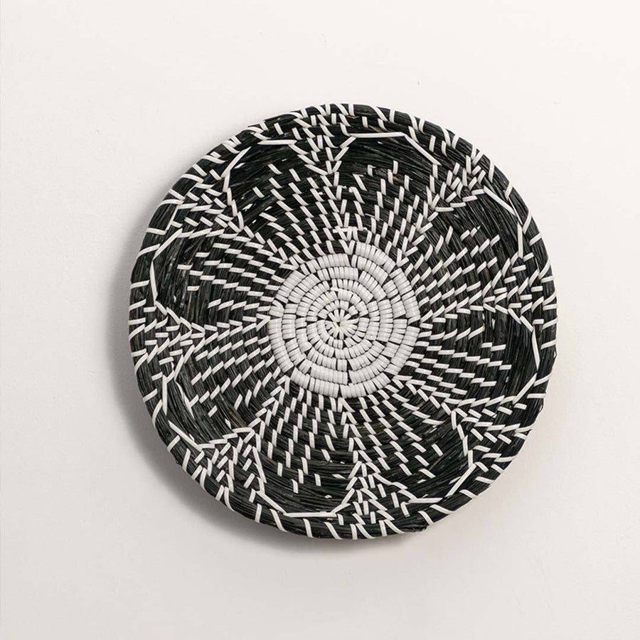 Round handcrafted black cream woven seagrass wall basket with detailed multi petal flower pattern on white background image.