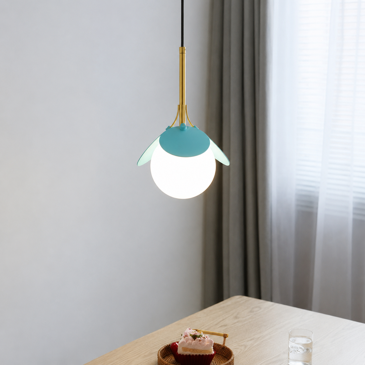 A modern pendant light hanging over a wooden table in a softly lit room. The lamp has a white spherical globe shade, a turquoise-blue top cap, and a gold-toned metal stem and side supports.