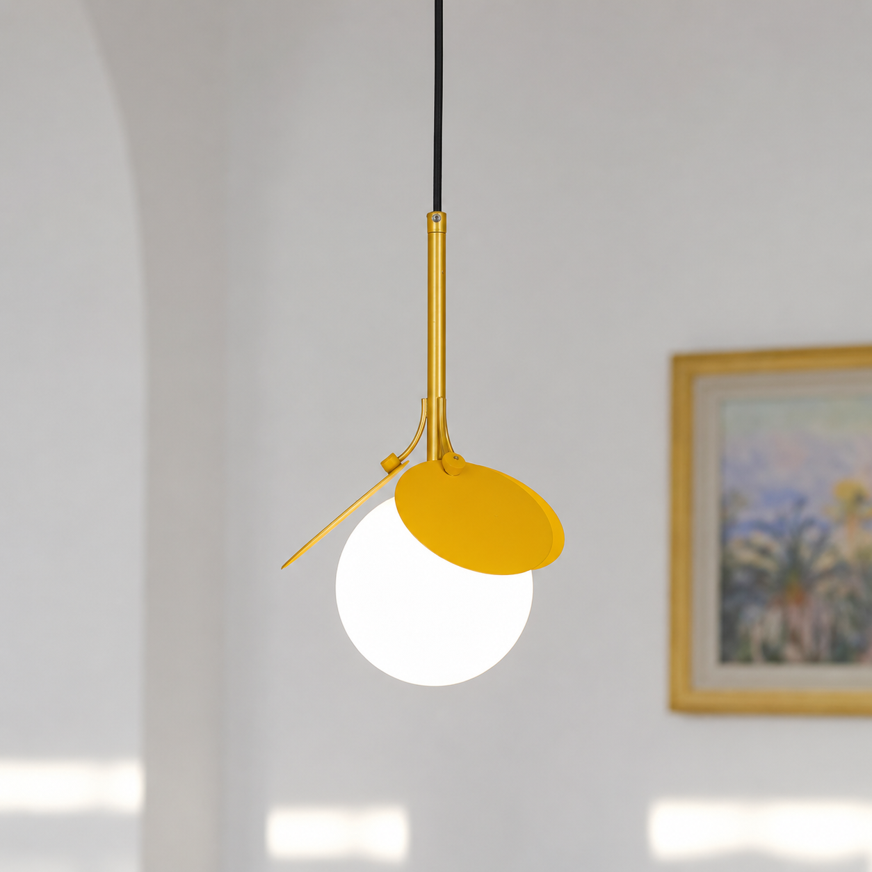 A modern pendant light with a matte yellow shade, a white globe bulb, and a gold metal stem suspended by a black cord in a bright, minimalist room.