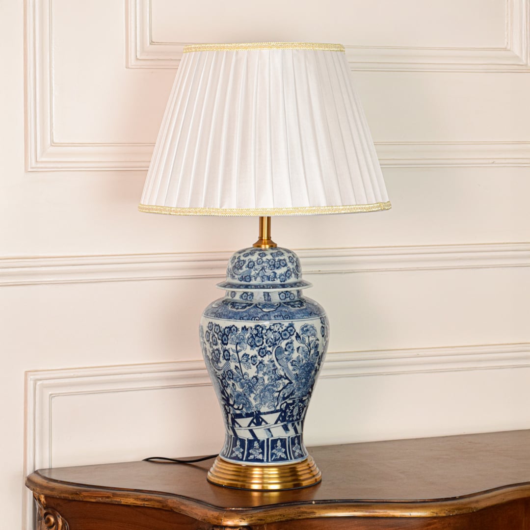 Elegant blue white ceramic table lamp with ornate floral design gold metal base and pleated fabric shade on off white background.
