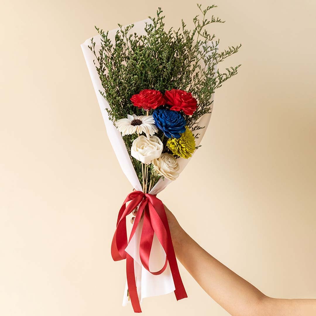Main lifestyle image showing preserved multicolor sola flower bouquet with green limonium grass wrapped in white paper and red ribbon held by hand in elegant setting.