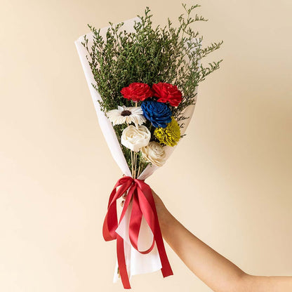 Main lifestyle image showing preserved multicolor sola flower bouquet with green limonium grass wrapped in white paper and red ribbon held by hand in elegant setting.