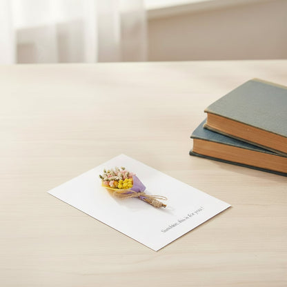 A handmade white greeting card with a mini yellow dried flower bouquet resting on a table next to stacked vintage books.