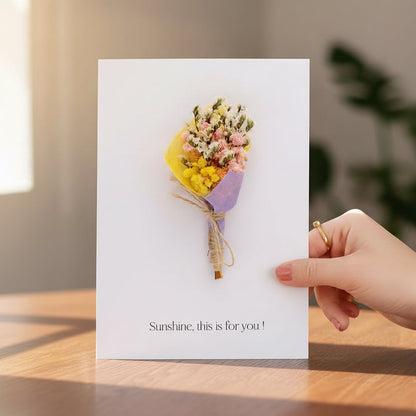 Hand holding a white greeting card featuring a colorful mini dried flower bouquet resting on a sunlit wooden tabletop.