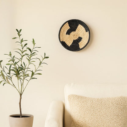 A handwoven natural and black rattan wall decor plate styled above a marble table with a tall black vase and geometric sculpture creating warm modern home decor vibes.