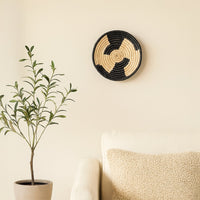 A handwoven natural and black rattan wall decor plate styled above a marble table with a tall black vase and geometric sculpture creating warm modern home decor vibes.