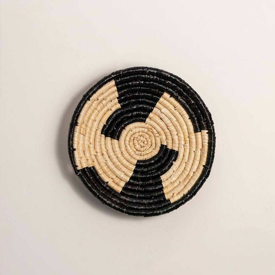 Round handcrafted black and cream woven seagrass wall basket with geometric swirl block pattern on white background image.