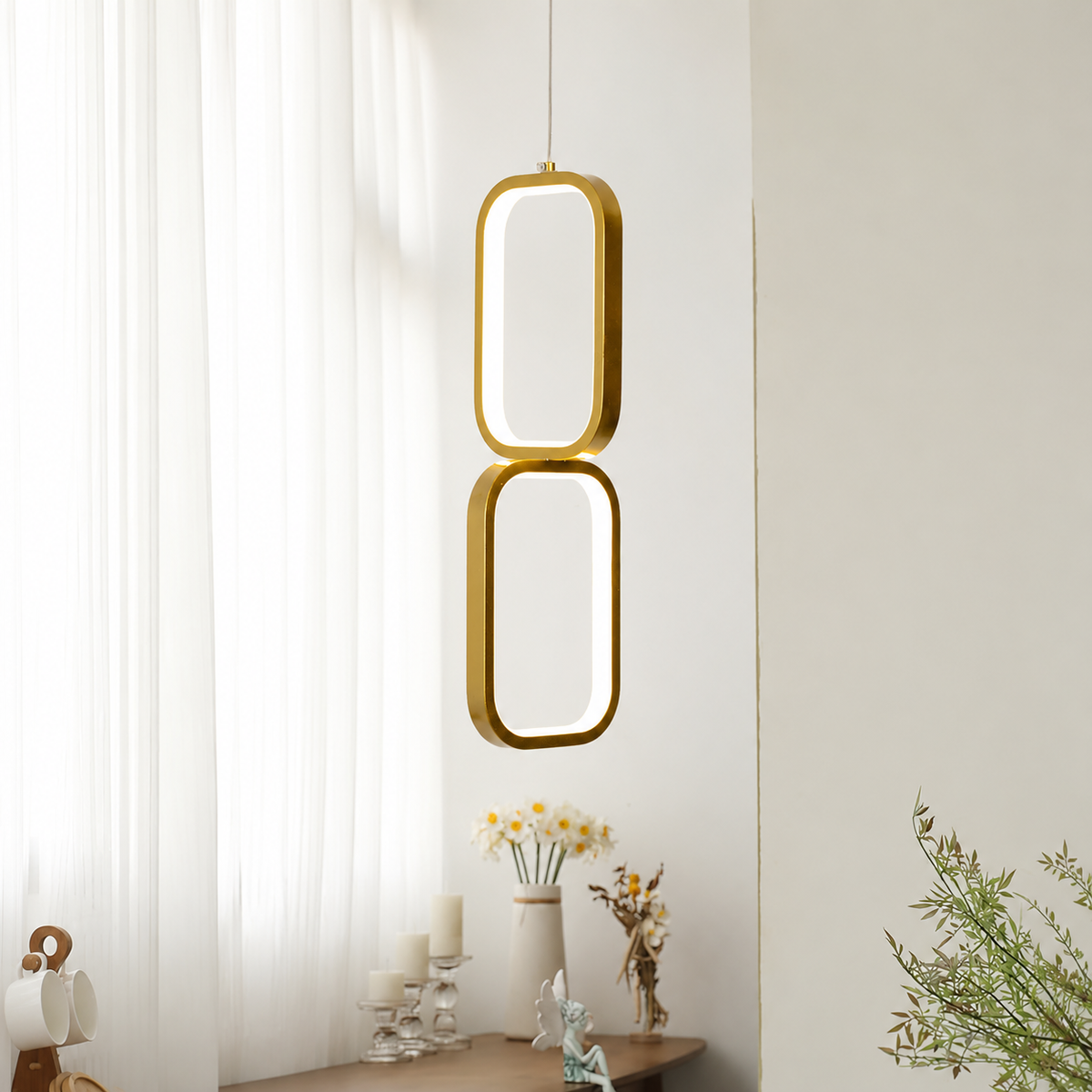 A modern two-ring pendant light with a brushed gold finish and a soft white glow, hanging in a bright, minimalist room with sheer white curtains and a wooden table below.