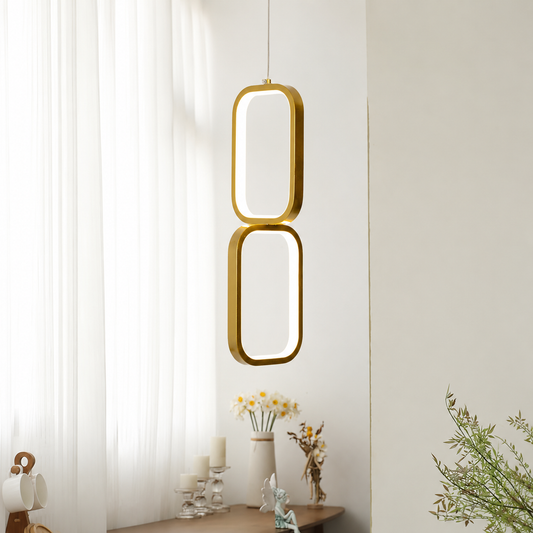 A modern two-ring pendant light with a brushed gold finish and a soft white glow, hanging in a bright, minimalist room with sheer white curtains and a wooden table below.