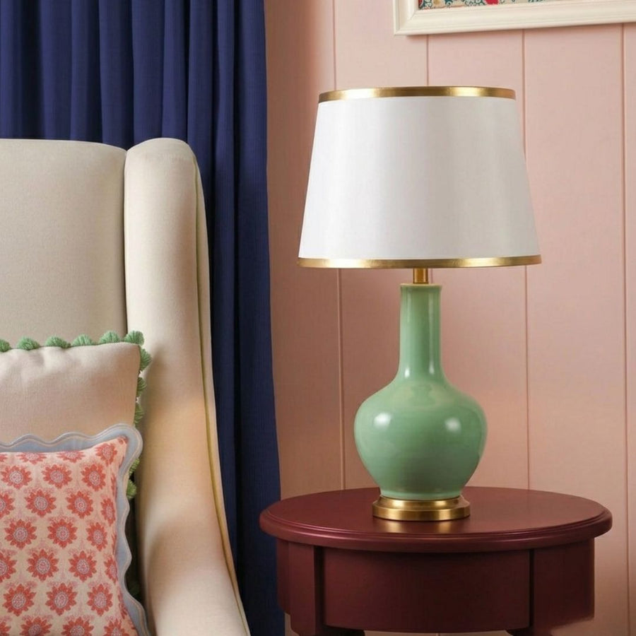 A table lamp with a glossy mint green ceramic base, a white drum shade, and gold trim, styled on a dark wood side table next to a cream armchair.
