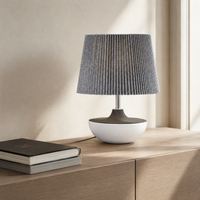A modern table lamp with a gray pleated fabric shade, a dark wood-toned middle section, and a white ceramic bowl base, styled on a light wood sideboard next to a stack of books.