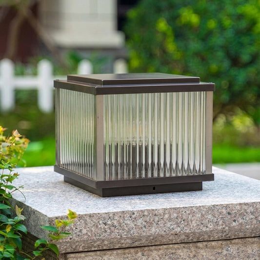 Outdoor pillar light with rectangular metal frame and ribbed glass shade installed on stone base in garden setting for landscape lighting.