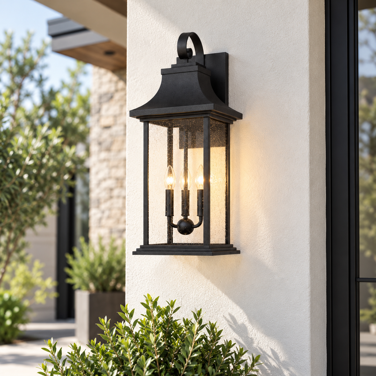 Black outdoor wall sconce with a warm glowing light, mounted on a beige stucco exterior wall with green plants in the foreground.