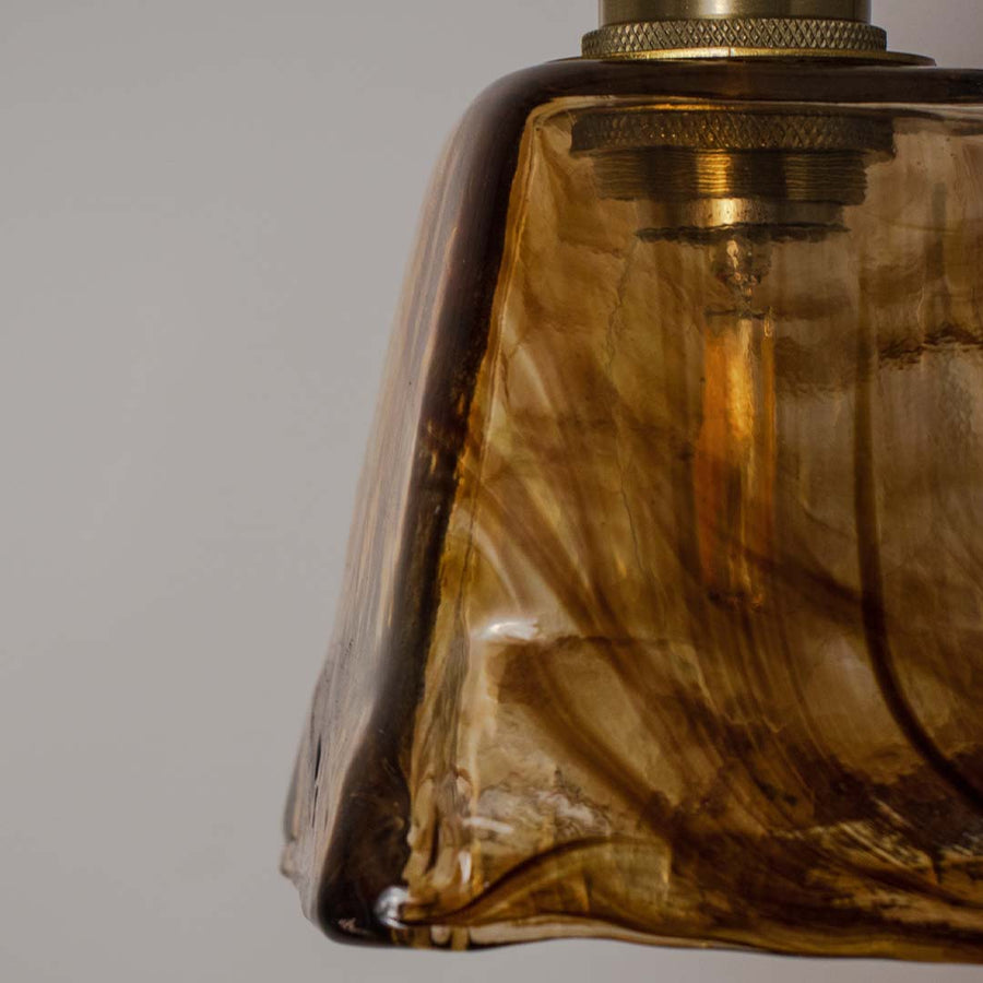 Closeup amber glass pendant light with smooth curved shade and gold fitting placed against neutral wall background highlighting rich warm lighting design.