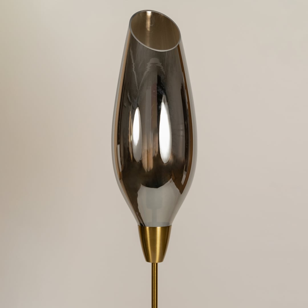 Close up of a floor lamp with an elongated smoky grey glass shade on a slim gold stand showing the bulb softly visible inside.