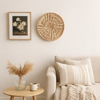 A handwoven natural rattan wall decor plate styled above a beige sofa with a wooden side table glass vase and soft neutral accents creating warm cozy home decor vibes.