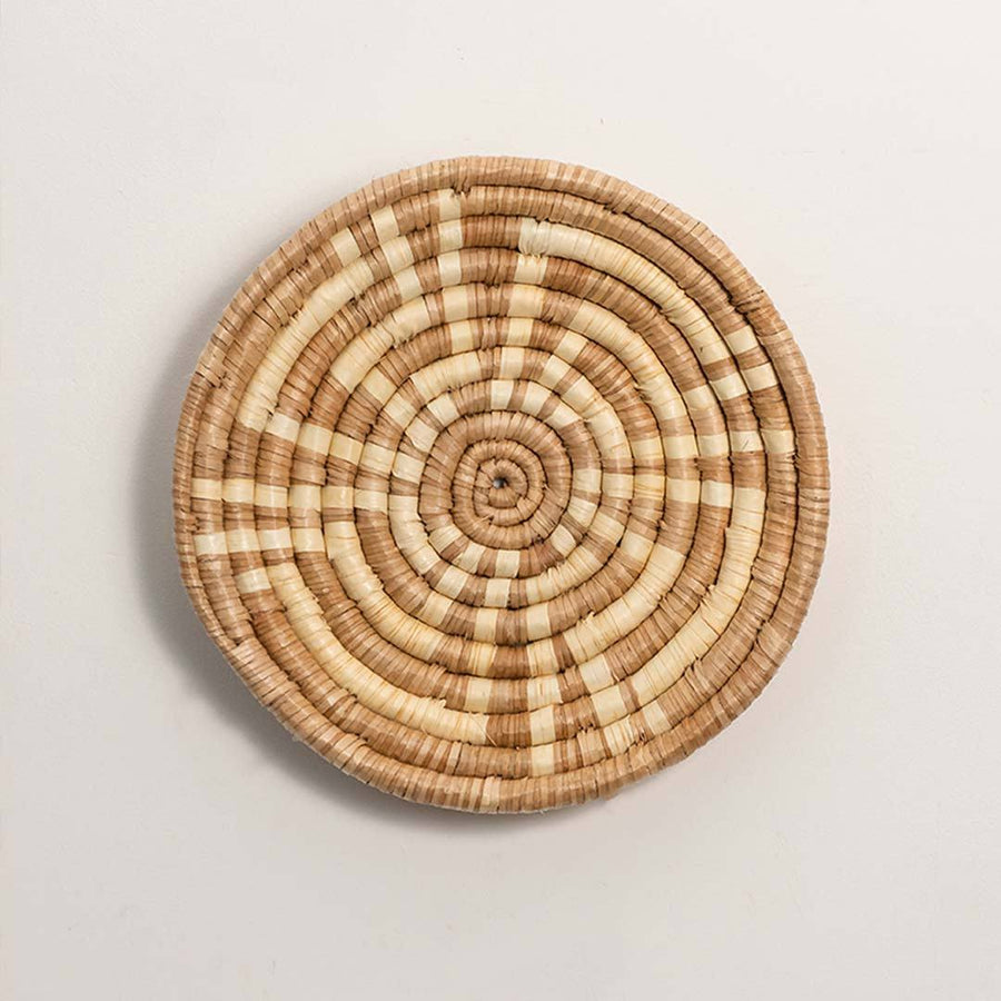 Round handcrafted natural brown cream woven seagrass wall basket with concentric spiral pattern on white background image.