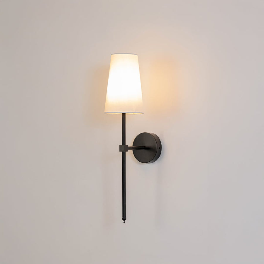 Illuminated white fabric drum shade wall light with tall black metal rod emitting warm ambient glow on white background.