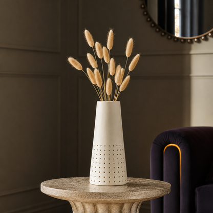 A white ceramic vase with a perforated lower section, holding dried bunny tail grass, styled on a round stone side table against a dark paneled wall.