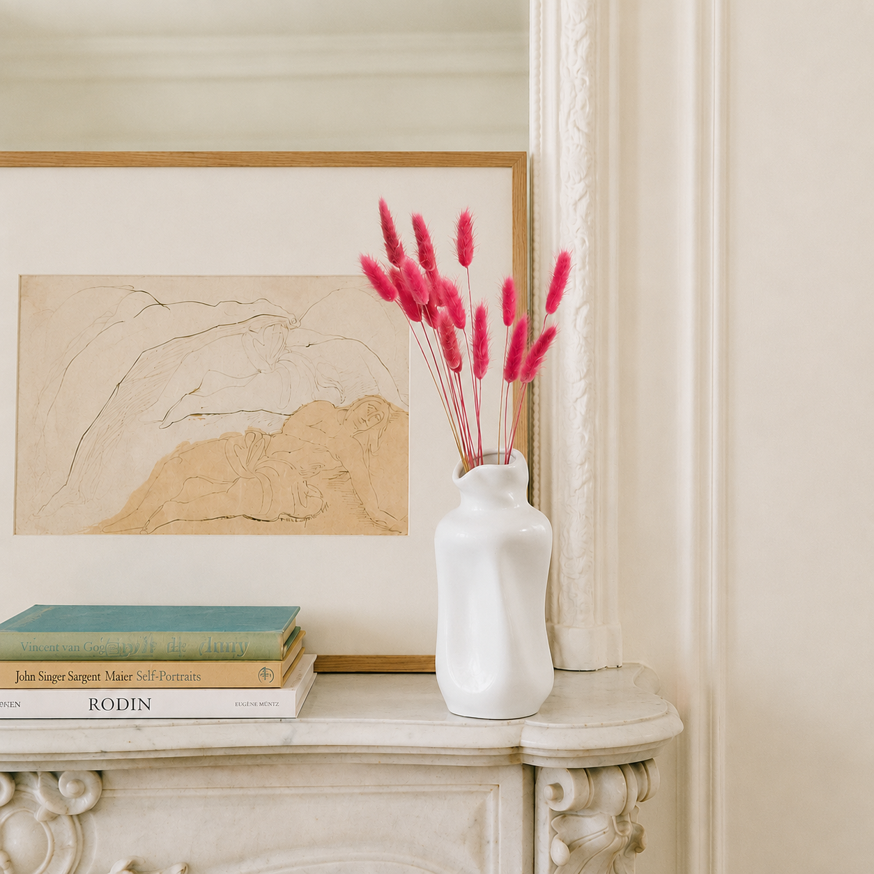 A white ceramic vase filled with bright pink bunny tail grass stems sits on a cream-colored mantelpiece next to a framed abstract landscape print and stacked books.