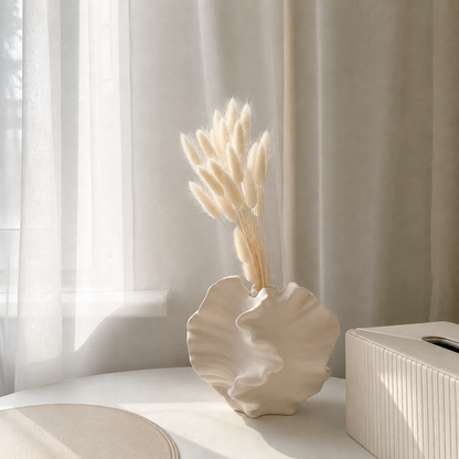 A cream-colored ceramic vase with a scalloped edge, holding dried bunny tail grass, styled on a white table next to a ribbed white tissue box box in front of sheer curtains.