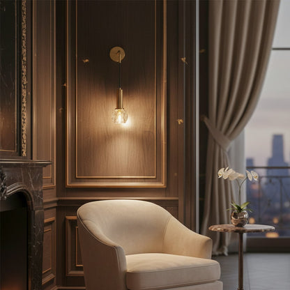 Lifestyle image of gold metal wall light with clear glass shade mounted beside lounge chair adding warm ambient lighting.