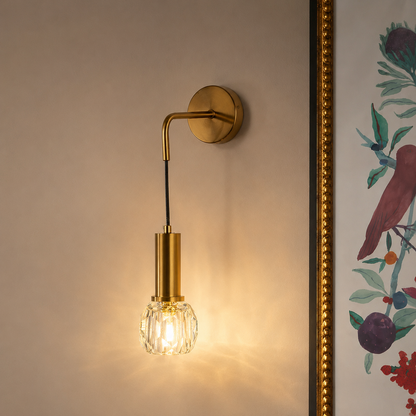 A modern wall sconce with a brushed gold metal mount and arm, a black hanging cord, and a faceted clear glass shade glowing with warm light, shown mounted on a beige wall next to a framed botanical print.
