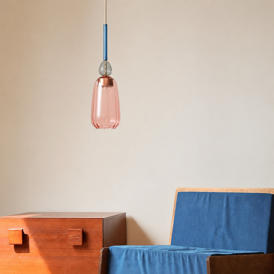 A modern pendant light with a translucent pink glass shade and a blue cord, hanging in a minimalist room above a wooden side table and a blue upholstered chair.