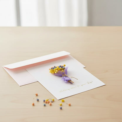 A handmade greeting card featuring a mini yellow and purple dried flower bouquet resting by a pink envelope on a desk.