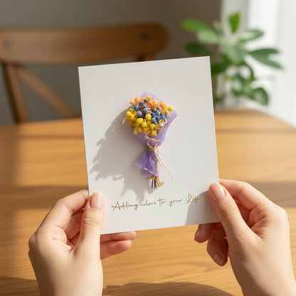 Hands holding a handmade greeting card with a mini yellow and purple dried flower bouquet in a warm sunlit living room.