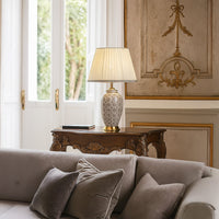 Elegant grey patterned table lamp with gold base placed on a carved wooden console in a classic living room with soft natural daylight.