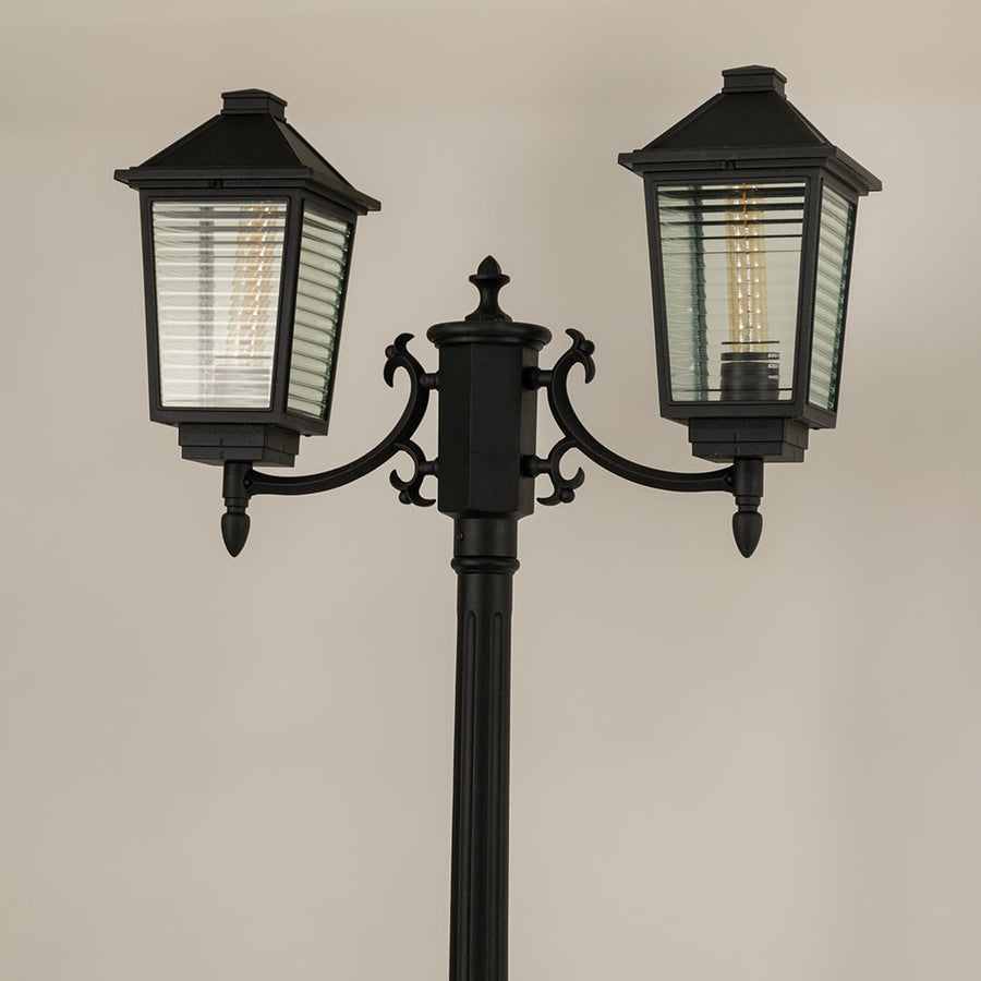 Closeup of black metal dual lantern lamp post showing ribbed glass panels and elegant curved arms perfect for outdoor pathway garden and porch Decor.
