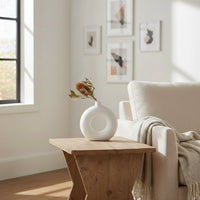 Modern white ceramic donut vase placed on wooden side table beside beige sofa enhancing minimalist living room decor with dried floral styling.