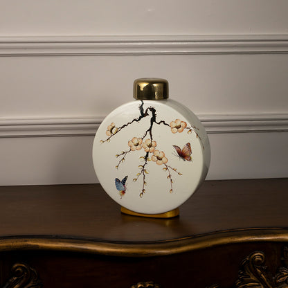 Round ceramic vase with bird and branch artwork and gold neck placed on wooden table against neutral wall background showcasing elegant decorative home styling.