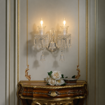 An ornate two-light crystal wall sconce mounted on a beige paneled wall above a carved gold console table with white flowers.