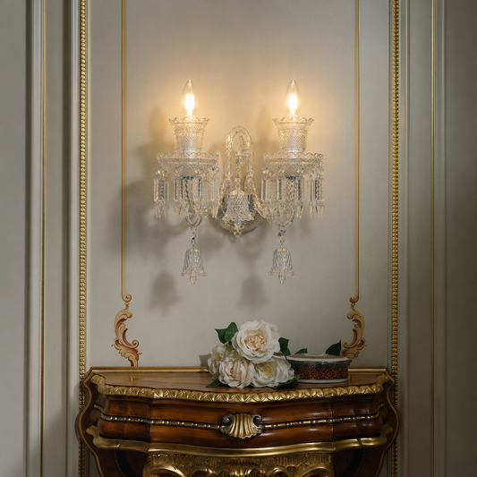 An ornate two-light crystal wall sconce mounted on a beige paneled wall above a carved gold console table with white flowers.